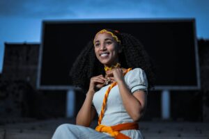 a woman in white top with yellow and brown headband and choker